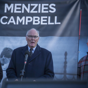 Menzies Campbell speaking at a political rally. Wearing a navy blue suit and glasses. Dark background with political slogans in bold letters