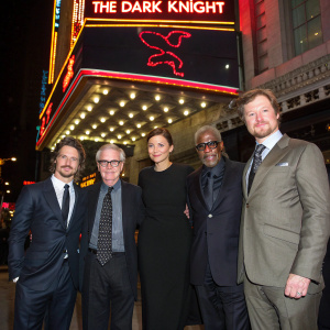 Christian Bale, Gary Oldman, Michael Caine, Maggie Gyllenhaal, Morgan Freeman, and Christopher Nolan at a film festival. Wearing suits, standing in front of neon-lit billboard featuring 'The Dark Knight' movie title and studio logos