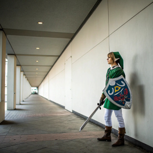 A fan dressed as Link from 'The Legend of Zelda' standing alone in a quiet hallway. Minimalistic white wall backdrop.