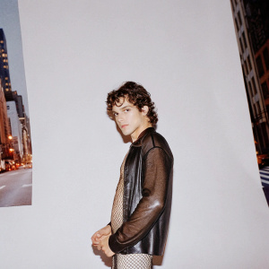 Austin Mahone, Hispanic male, with curly hair. Wearing a black leather jacket with mesh detailing. Posing for an advertisement campaign in a busy street, surrounded by vibrant city lights.