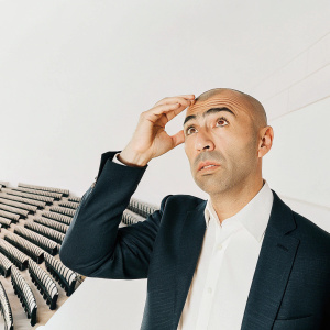 Roberto Di Matteo, bald, wearing a blue suit and white shirt, stands in a stadium filled with seats. He looks worried.