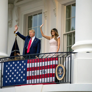 President Trump and First Lady Melania Trump standing on the balcony of the White House. They wave to the crowd. American flag and White House windows in the background.