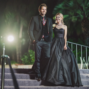 Chad Michael Murray and Kenzie Dalton at a glamorous gala. Chad, dressed in a sharp black tuxedo with a vibrant patterned tie, stands beside Kenzie, adorned in a stunning dark floor-length gown. They share a warm smile for the camera. Dimly lit setting with spotlight.