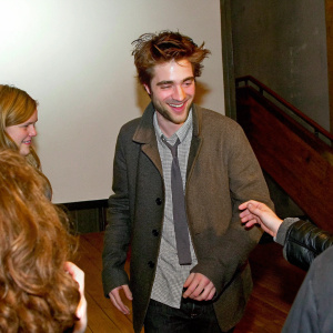 Robert Pattinson, with a joyful smile, in a stylish yet casual outfit. Surrounded by fans at an indoor event. Simple, modern backdrop.