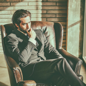 Jake Gyllenhaal, seated in a plush leather chair, wears a dark suit with a neatly styled haircut. Thoughtful expression under warm, soft lighting. Brick wall background blurred