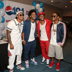 B2K members Raz-B, Omarion, Lil Fizz, and J-Boog at a promotional event. The group is dressed casually with sporty outfits. The background features a banner with balloons and a table with a microphone and drinks.