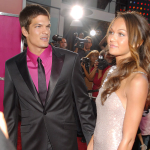 Rick Malambri and Sharni Vinson at a glitzy Hollywood premiere. Rick in a sleek black tuxedo with purple shirt, Sharni in a sparkling silver dress. Crowd and flash photography visible.