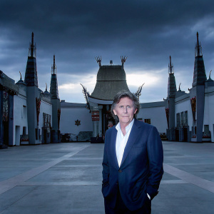 Gabriel Byrne, in a navy blue suit and crisp white shirt, stands outside the TCL Chinese Theatre in Hollywood.