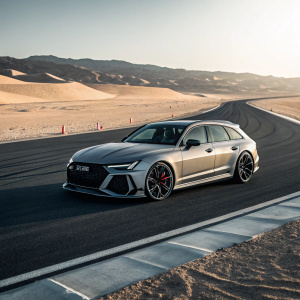High-performance Audi RS6 Avant parked on a gleaming asphalt track amidst a sprawling desert vista. The car's sleek design, bold grille, and aerodynamic silhouette are accentuated by dramatic lighting.