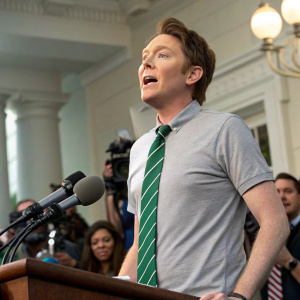 Clay Aiken, standing at a press conference. Wearing a gray t-shirt, green striped tie. Expressive face, dynamic pose.