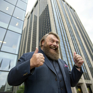 Brian Blessed, with his iconic beard and broad smile, making a thumbs-up gesture outside a corporate headquarters building.