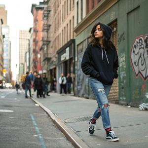 Camila Cabello on the streets of New York City. She's casually dressed in a black hoodie, ripped jeans, and sneakers. Graffiti-covered walls and bustling city life in the background.