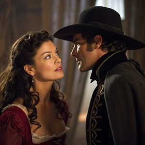 In a dimly lit scene from The Legend of Zorro, Catherine Zeta-Jones appears playful and curious, while Antonio Banderas, dressed in a Spanish-style outfit, whispers nearby. The background is blurred with a curtain-like texture.