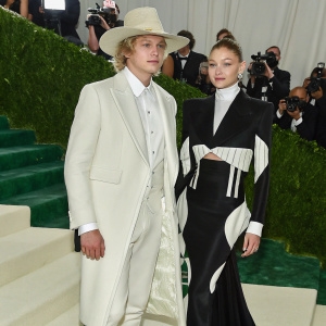 Cody Simpson and Gigi Hadid at the Met Gala. Cody in a black tuxedo, crisp white shirt, and beige hat. Gigi in a sleek black gown with geometric cutouts. The backdrop is a bustling red carpet with flashing cameras.