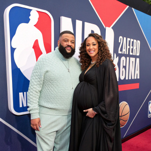 DJ Khaled and Nicole Tuck at the NBA All-Star Game. DJ Khaled in a light grey sweater, Nicole Tuck in a flowing black gown. The backdrop features the NBA logo.