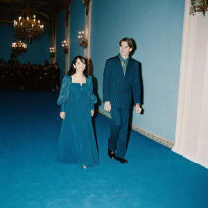 Devika Bhise and Jeremy Irons smiling at a glamorous charity gala. Dimly lit ballroom with chandeliers and a crowd of well-dressed attendees.