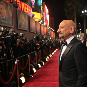 Sir Ben Kingsley at a glitzy Hollywood gala. Black tuxedo, slight smile. Crowded red carpet, flashing cameras, paparazzi, and vibrant movie posters surrounding him.