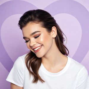Hailee Steinfeld, featuring a close-up shot of her smiling face, makeup enhanced. Dressed in a simple white t-shirt. Heart-shaped purple backdrop.