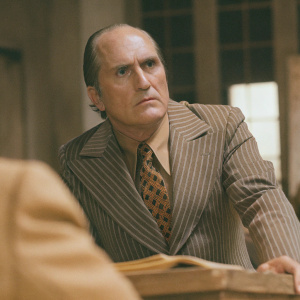 Robert Duvall as Frank Hackett from Network. Serious expression, in a pinstriped suit with a bold patterned tie. Shot in dimly lit room with blurred background.