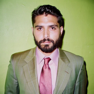 retrato mugshot de hombre latino con barba corta vestido de traje formal en fondo de un color azul