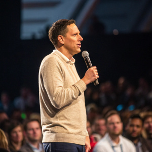Peter Thiel, in a casual beige sweater, giving a keynote speech. Crowd in background, blurry, attending a tech conference.