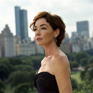 Professional portrait of Julianne Nicholson, actress with short brown hair. Serious expression, close-fitting dark strapless dress. Shot in busy city park, blurred buildings and skyline in background. Natural lighting, soft focus on actress, highlights features.