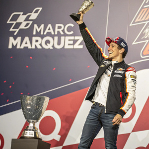 Marc Marquez, posing with a trophy. Dynamic pose, celebrating a victory. Spanish text and logos in the background.