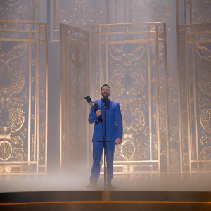 Justin Timberlake grinning on the iHeartRadio Music Awards stage, clad in a vibrant blue suit, holding a sleek, dark award. Luxurious, gold-embroidered backdrop