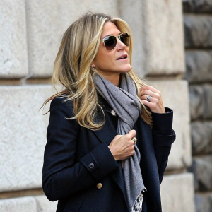 Jennifer Aniston, blonde hair, sunglasses, and scarf, adjusting her coat. Stone wall background, candid photo.