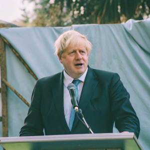 Boris Johnson, in a dark suit and blue tie, speaking at a press conference. The backdrop is a solid blue color.