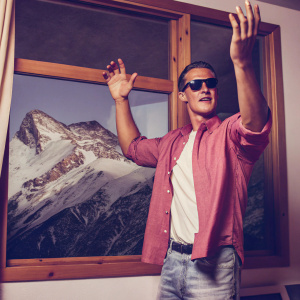 Michael Schumacher in casual attire, red shirt and sunglasses, poses with hands up. Snowy mountains backdrop