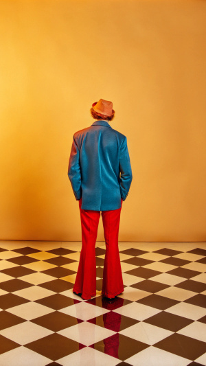 A mid-aged individual photographed from a full posterior view in a distinctive 1980s aesthetic, standing on a black and white chessboard-patterned floor. Walls are pure white, creating a stark backdrop. The subject wears an electric blue ensemble characteristic of 1980s fashion - potentially with sharp shoulder lines, geometric cuts, or bold textures. The image captures the era's visual language: high contrast, slightly grainy film quality, with a color palette and styling that evokes the bold, synthetic visual culture of the decade. Lighting has a slightly harsh, direct quality typical of 1980s photography.