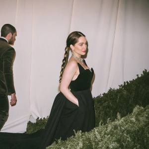 Emilia Clarke, adorned in a sleek black evening gown with halter neck, graces a gala event. Sophisticated makeup and a braid with gold accents highlight her striking appearance. A plain white backdrop.