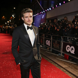 Richard Madden, clad in a formal black tuxedo, poses confidently at the GQ Men of the Year Awards. Glittering red carpet with flashing cameras