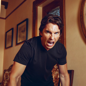 Christian Bale in an intense close-up. Dark brown eyes locked with the camera. Fierce expression. Black t-shirt hugs his physique. Warm, earthy background tones.