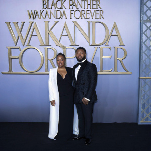 Zinzi Evans and Ryan Coogler attend premiere. Dressed in elegant black and white formal attire. Standing in front of large gold-patterned backdrop featuring 'Black Panther: Wakanda Forever' title