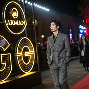 Park Hyung-Sik at a high fashion event, dressed in a tailored Armani suit. Standing against a glowing, neon-lit backdrop featuring interlocking G logos, in a bustling night event.