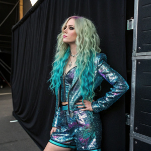 Avril Lavigne in a promotional shoot. Shiny outfit with holographic design, turquoise curls. Poses in front of black backdrop with brand logos.