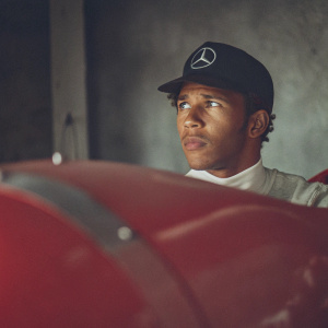 Close-up shot of Lewis Hamilton, intense stare, wearing black Mercedes AMG cap. Sparse background.