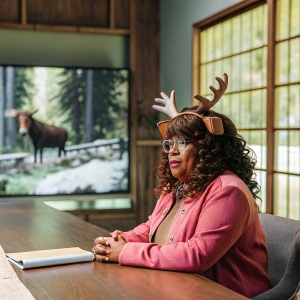 Sherri Shepherd as Anne Flatch from Trial and Error: Lady Killer. Curly brown hair, pink jacket, brown antler headpiece. Sitting at a wooden table in a modern, brightly lit studio with a large TV displaying moose imagery behind her.
