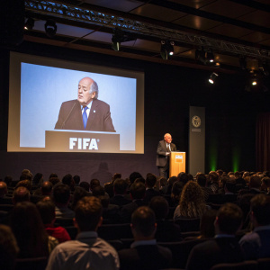 Sepp Blatter addressing a crowded auditorium during a FIFA press conference. Projector screen behind him displaying FIFA logo.
