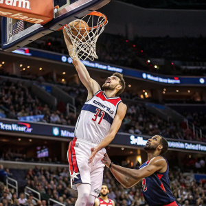 Tomas Satoransky, dressed in Wizards jersey, leaping high for a slam dunk during a heated basketball match. His rival, clad in opposing team's uniform, reaches out to block the shot. Crowd roars in excitement.