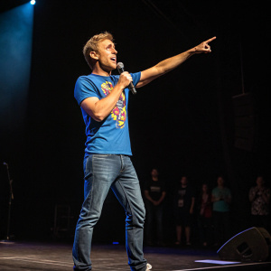 Russell Howard, comedian, in energetic performance stance. Wearing blue graphic t-shirt, on stage with spotlight