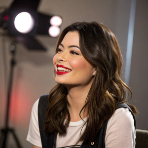 Miranda Cosgrove, smiling in an interview setting. Wearing casual outfit with bold red lipstick. Studio lights casting soft shadows.