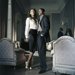 Nick Cannon and Selita Ebanks pose at an upscale charity gala. Selita wears a loose-fitting white blouse and black skirt. Nick sports a dark suit and striped shirt. They stand in a room filled with elegant furniture and distinguished guests.