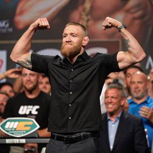 Conor McGregor at a boxing event. No beard, wearing a black shirt, flexing muscles. The background is filled with cheering fans.