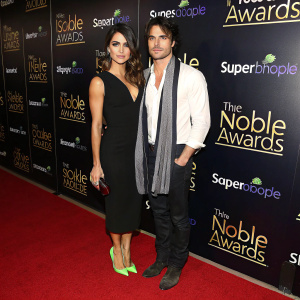 Nikki Reed and Ian Somerhalder at the Noble Awards gala. Nikki in a black dress with green heels, Ian in a white shirt and scarf. They stand on the red carpet against a backdrop of event logos and bright lights.
