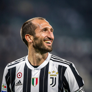 Giorgio Chiellini, smiling widely. Dressed in Juventus football jersey with team sponsors' logos. Blurry stadium background