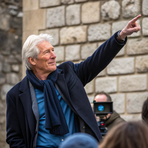 Richard Gere, actor, pointing confidently. Wearing dark jacket, blue shirt, and dark scarf. Candid moment at an event with stone wall backdrop.