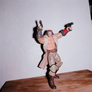 High-definition photo of a detailed, one-quarter scale Hellboy action figure. The miniature is captured mid-action, showing fierce determination. The character, featuring demonic features and adorned with a belt, armor, and weapons, is set against a dark, textured background.